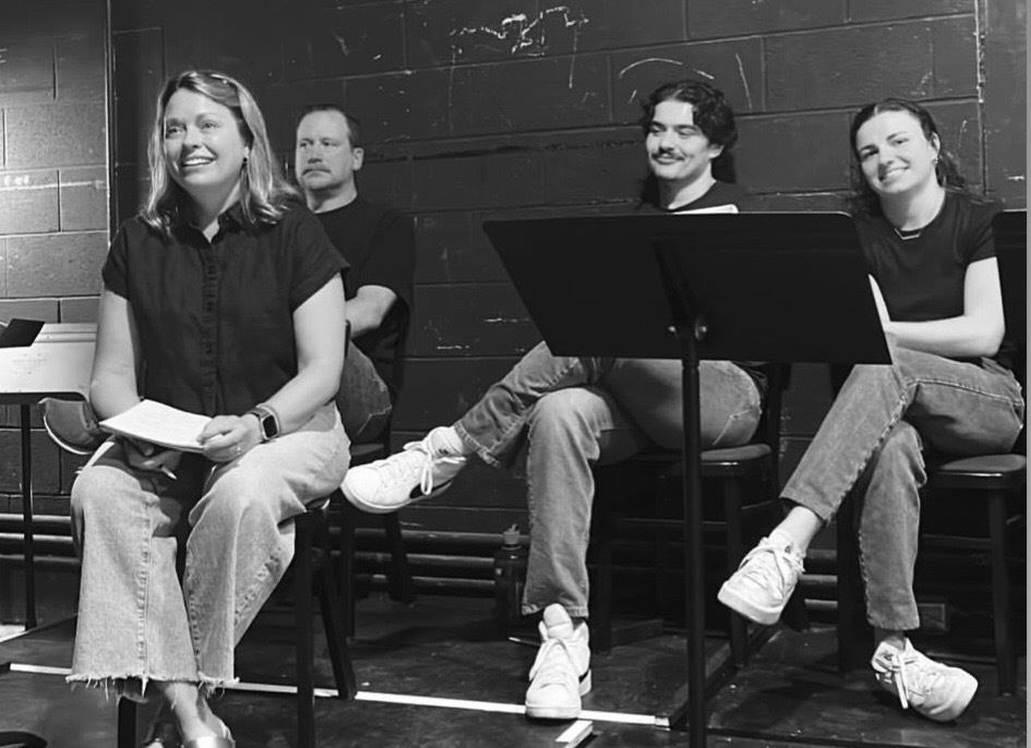 Cast members seated on a black box stage between scenes