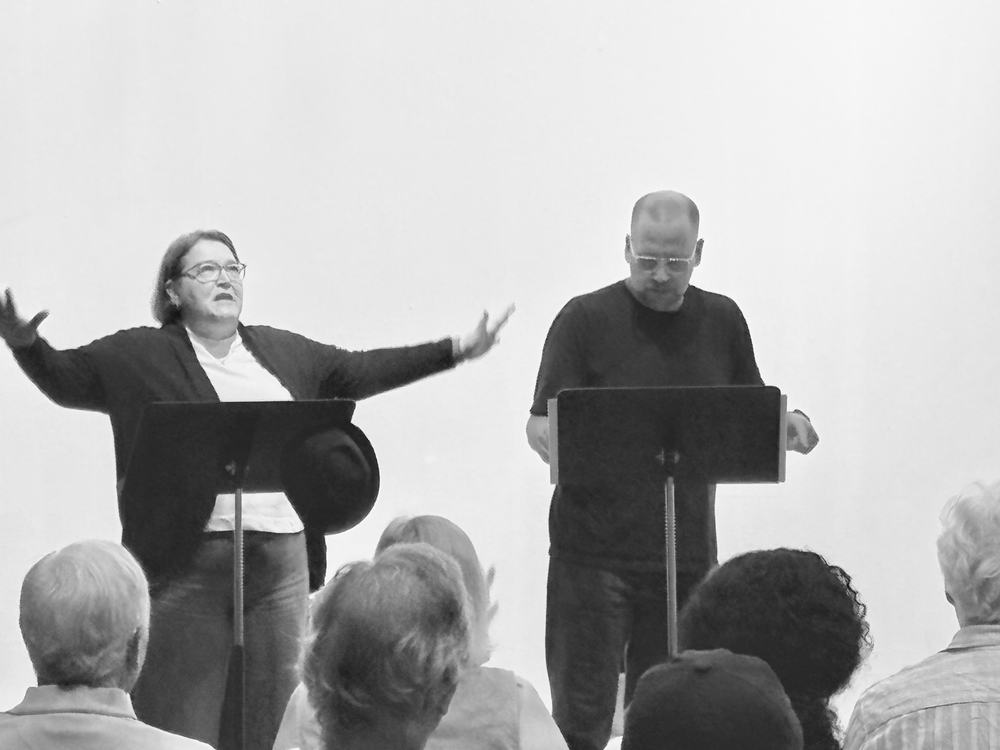An actor with arms outstretched mid-scene beside a second reader